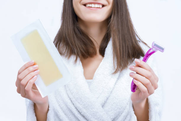 Woman in white robe holding wax strip and razor, smiling